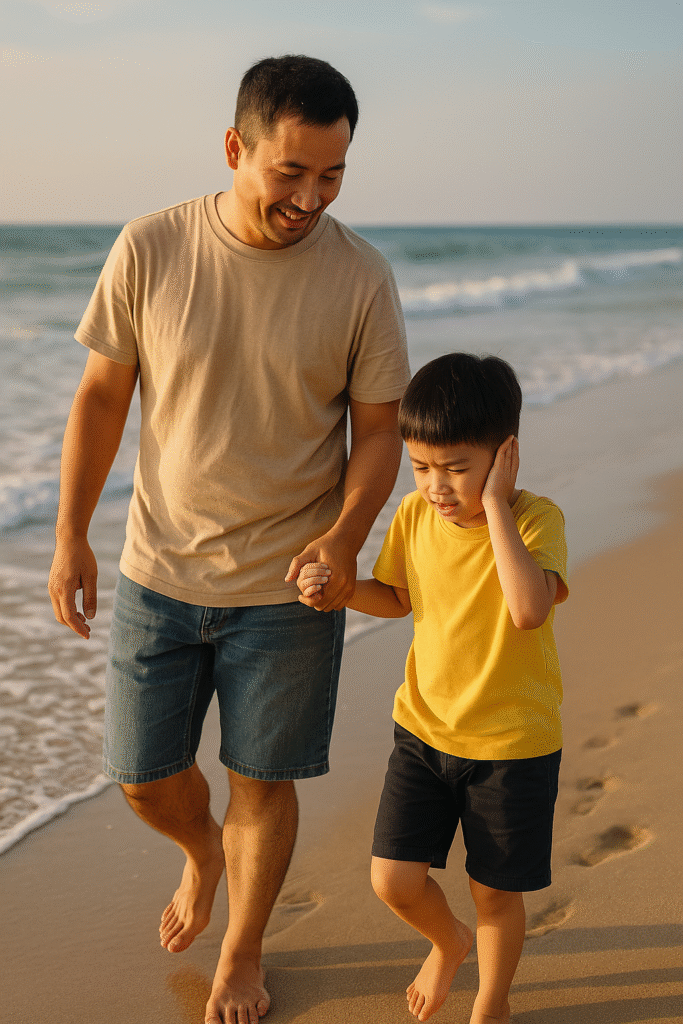 Người cha dắt con tự kỷ đi dạo trên bãi biển lúc hoàng hôn, bé che tai vì nhạy cảm âm thanh.